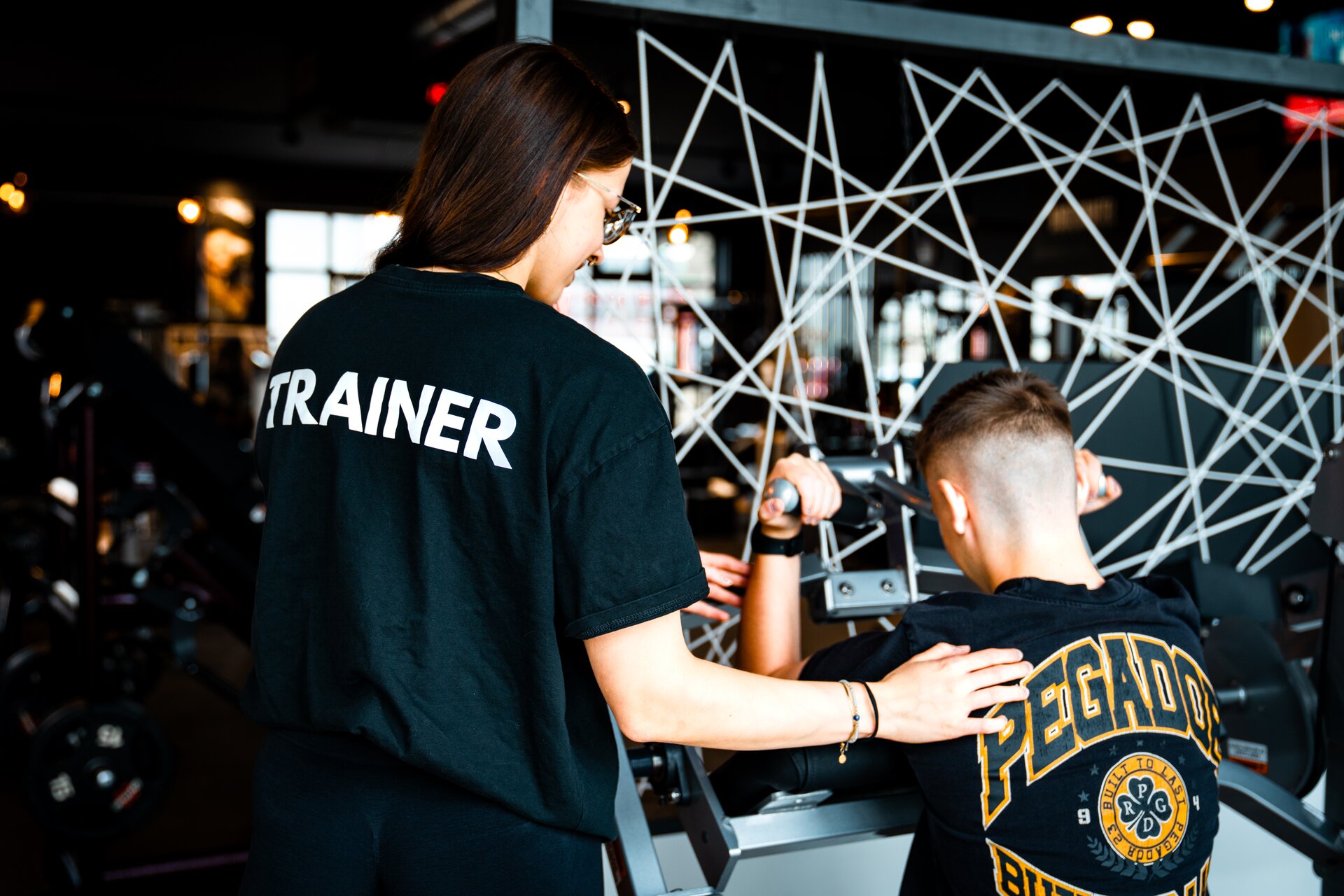 Trainerin mit TRAINER-Shirt betreut Mitglied an der Maschine im Fitnessstudio Bitburg GymOne 24/7